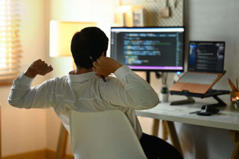 Back view of male programmer stretching arms and relaxing on chair at his workplace.