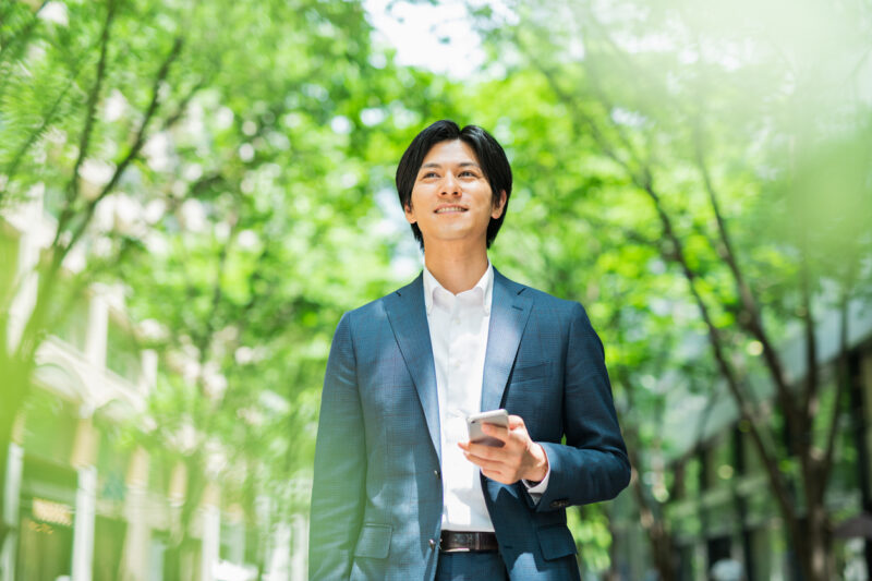 Young businessman walking down a street with street trees, looking at his smartphone.