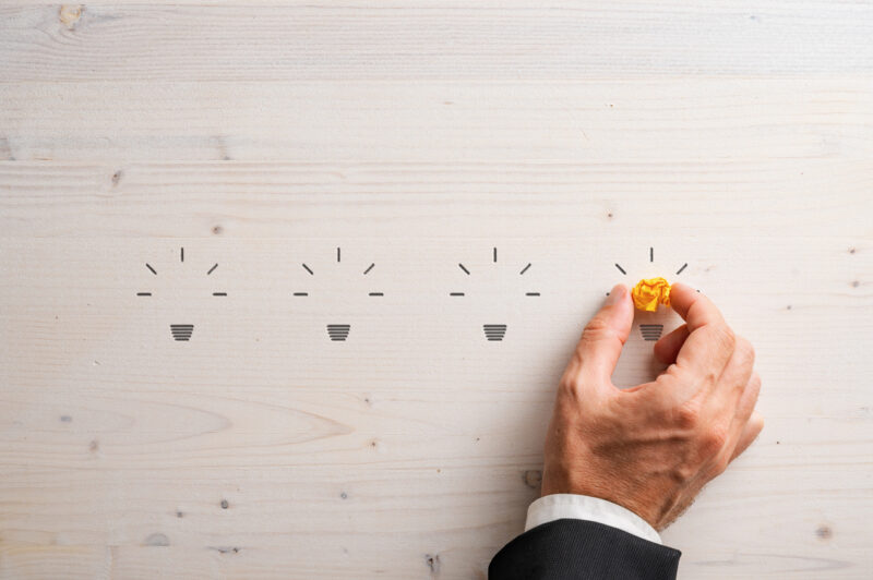 Silhouettes of four light bulb shapes carved into wooden background with a hand of a businessman placing smashed yellow paper in the fourth one. Conceptual image of idea and innovation.