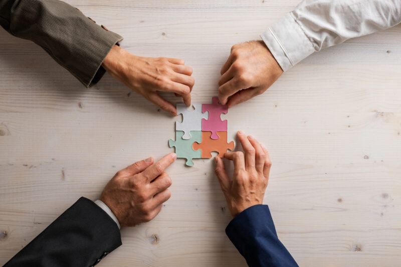 Hands of four businesspeople joining matching puzzle pieces of various pastel colors over a wooden background.